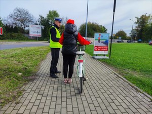 Zdjęcie kolorowe przedstawia umundurowanego policjanta, który stoi obok kobiety, która pcha rower po chodniku.