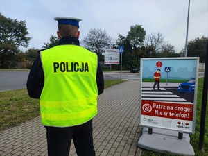 Zdjęcie kolorowe przedstawia umundurowanego policjanta ruchu drogowego obok kolorowego plakatu informacyjnego.