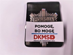 Zdjęcie kolorowe: policyjna odznaka, a pod spodem hasło fundacji Pomogę, bo mogę DKMS