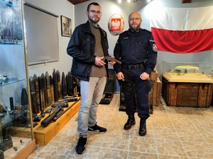 Zdjęcie kolorowe przedstawia umundurowanego policjanta obok mężczyzny, który trzyma w rękach zabytkowy pistolet.