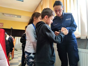 Zdjęcie kolorowe przedstawia umundurowaną policjantkę, która pokazuje dziecku kajdanki.