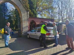 Zdjęcie kolorowe przedstawia umundurowanego policjanta, która zakłada kobiecie opaskę odblaskową.