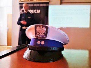 Zdjęcie kolorowe przedstawia na pierwszym planie policyjną czapkę ruchu drogowego, w tle widoczny policjant.