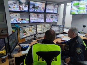 Zdjęcie kolorowe przedstawia umundurowanego policjanta i strażnika miejskiego przed ekranami monitoringu miejskiego.