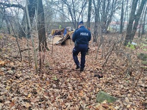 Zdjęcie przedstawia policjanta idącego przez las. Z tyłu widoczny namiot.