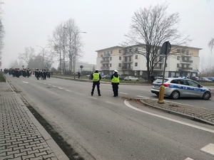 Zdjęcie przedstawia dwóch umundurowanych policjantów, radiowóz. Z tyłu widoczna orkiestra dęta.