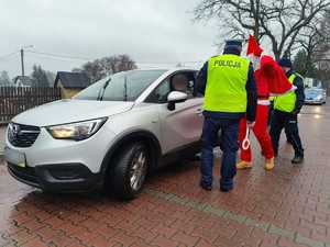 Zdjęcie przedstawia policjantów i Mikołaja podczas kontroli drogowej. Wszyscy stoją obok srebrnego samochodu.