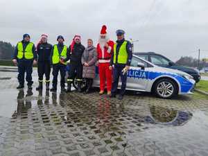 Zdjęcie przedstawia policjantów, strażaków, Mikołaja i kobietę. Z tyłu widoczny radiowóz policyjny.