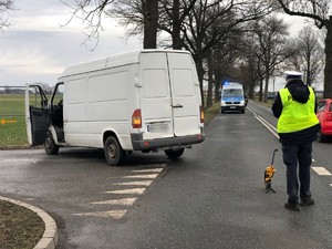 Na zdjęciu widoczny policjant podczas czynności służbowych, obok stoi samochód typu bus.