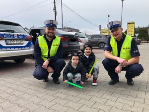 Zdjęcie przedstawia umundurowanych policjantów ruchu drogowego wraz z dwójką małych chłopców którzy trzymają w rękach odblaski.