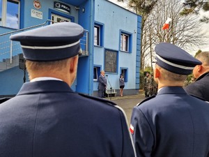 Zdjęcie przedstawia w oddali mężczyznę przed mikrofonem. Z przodu fotografii widoczni policjanci.