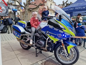 Zdjęcie przedstawia: dziecko które siedzi na policyjnym motocyklu. Z tyłu kobieta trzyma na rękach dziewczynkę. Wszyscy pozują do zdjęcia.