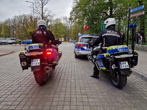 Zdjęcie przedstawia: dwa policyjne motocykle, które stoją tuż za oznakowanym policyjnym radiowozem.