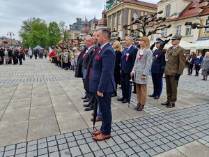 Zdjęcie przedstawia: uczestników uroczystości gdy stoją na baczność, z tyłu widoczny policjant i wojskowy podczas salutowania.