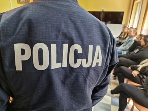 Zdjęcie przedstawia: napis Policja z policyjnego munduru, w tle widoczna młodzież.