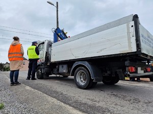 Zdjęcie przedstawia: policjanta przy samochodzie ciężarowym, z tyłu stoi kobieta w kamizelce odblaskowej.