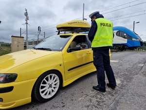 Zdjęcie przedstawia: policjanta podczas rozmowy z kierowcą samochodu osobowego, z tyłu widoczny przejazd kolejowy.