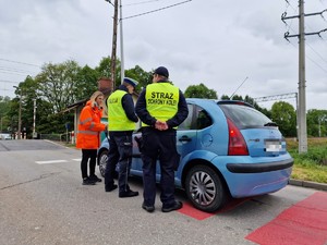 Zdjęcie przedstawia: mundurowi i kobieta w odblaskowej kurtce podczas rozmowy z kierowcą zatrzymanego samochodu osobowego.