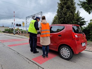 Zdjęcie przedstawia: umundurowanego policjanta ruchu drogowego i kobietę w kamizelce odblaskowej podczas rozmowy z kierowcą zatrzymanego pojazdu.