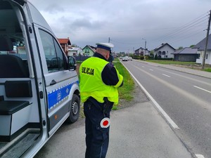 Zdjęcie przedstawia umundurowanego policjanta wydziału ruchu drogowego podczas pomiaru prędkości. Obok mundurowego stoi oznakowany radiowóz.