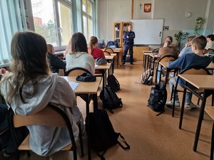 Zdjęcie przedstawia umundurowanego policjanta w sali szkolnej, gdy stoi przed siedzącymi w ławkach uczniami.