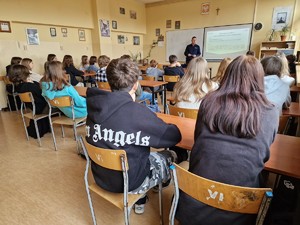 Zdjęcie przedstawia umundurowanego policjanta, który mówi do grupy młodych ludzi, którzy siedzą w ławkach szkolnych.