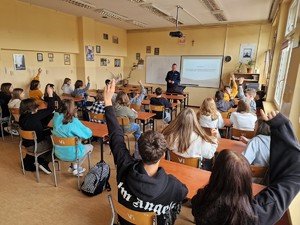 Zdjęcie przedstawia głoszących się uczniów w szkolnych ławkach, z tyłu widoczny policjant.