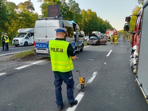 Zdjęcie przedstawia: z przodu widoczny policjant ruchu drogowego podczas czynności na miejscu wypadku drogowego. Z tyłu wszystkie służby ratunkowe.