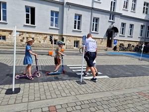 Zdjęcie przedstawia: troje dzieci i mężczyznę, który pokazuje jak zachować się bezpiecznie i prawidłowo na pasach