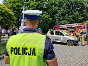 Zdjęcie przedstawia: umundurowanego policjanta w tle widać strażaków podczas  symulacji wypadku samochodowego