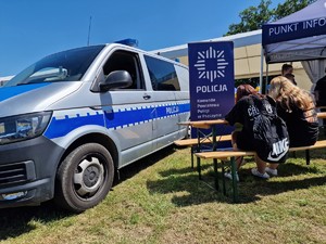 Zdjęcie przedstawia: policyjny radiowóz i osoby siedzące na ławce przy policyjnym stanowisku.