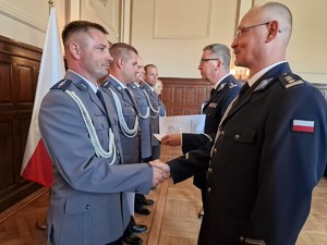 Zdjęcie przedstawia: rząd policjantów naprzeciwko których stoją ich przełożeni i wręczają nominacje na wyższy stopień policyjny oraz składają gratulacje.