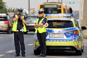 Na zdjęciu dwoje umundurowanych policjantów przy radiowozie. jeden z nich trzyma urządzenie do pomiaru prędkości.