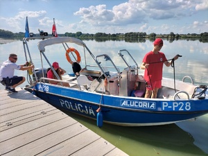 Zdjęcie przedstawia: policyjną łódź. Obok policjant i ratownicy.