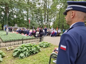 Zdjęcie przedstawia: umundurowanego policjanta podczas uroczystości. Z tyłu widoczni pozostali zgromadzeni.