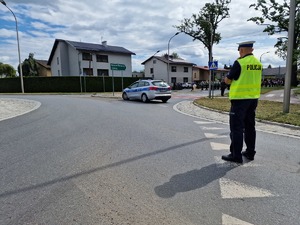 Zdjęcie przedstawia: policjanta blokującego ruch. Z tyłu widoczny radiowóz i pochód.