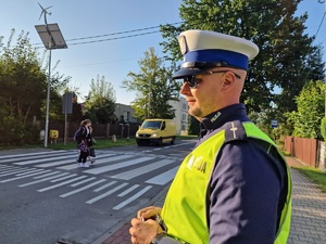 Zdjęcie przedstawia: umundurowany policjant stoi tuż obok przejścia dla pieszych. Z tyłu widoczna osoba dorosła z dzieckiem i samochód, który zatrzymał się ustępując im pierwszeństwa.