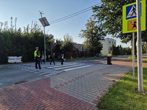 Zdjęcie przedstawia: mundurowy stoi na przejściu dla pieszych. Dzieci przechodzą przez jezdnie.