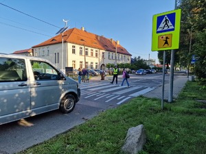 Zdjęcie przedstawia: policjanta, który stoi na przejściu dla pieszych podczas gdy dorośli i dzieci przechodzą.