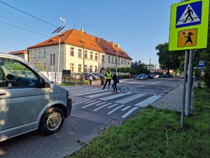 Zdjęcie przedstawia: dziecko na pasach pcha rower, obok stoi policjant.