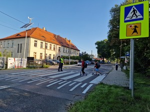 Zdjęcie przedstawia: policjanta na przejściu dla pieszych, dzieci które przechodzą pchając hulajnogi.