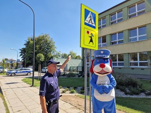 Zdjęcie przedstawia umundurowanego policjanta i policyjną maskotkę przy znaku drogowym informującym o przejściu dla pieszych w pobliżu szkoły.