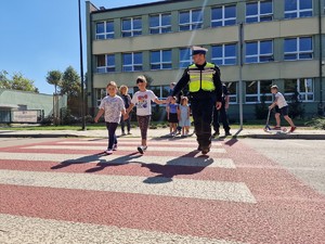 Zdjęcie przedstawia dwoje dzieci i policjant ruchu drogowego przechodzą przez pasy trzymając się za ręce.