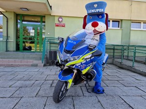 Zdjęcie przedstawia policyjną maskotkę na motocyklu pod szkołą.