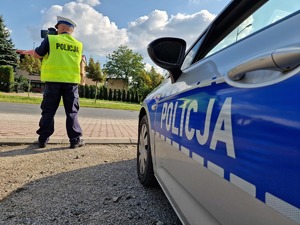 Zdjęcie przedstawia: umundurowany policjant podczas mierzenia prędkości, obok oznakowany radiowóz.