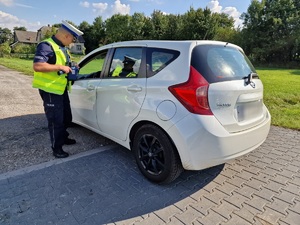 Zdjęcie przedstawia: policjanta stojącego przy białym samochodzie.