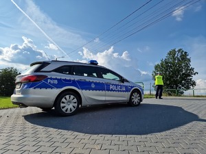 Zdjęcie przedstawia: oznakowany policyjny radiowóz, z tyłu widoczny policjant.