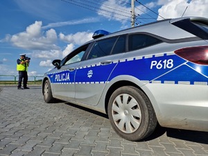 Zdjęcie przedstawia: umundurowanego policjanta obok oznakowanego radiowozu.