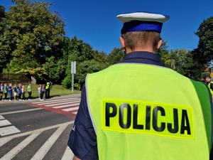 Zdjęcie przedstawia: policjanta ruchu drogowego, a po drugiej stronie jezdni innego policjanta wraz z dziećmi, które stoją przed przejściem dla pieszych.