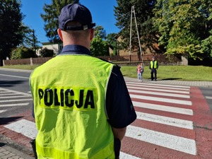 Zdjęcie przedstawia: umundurowanego policjanta, po drugiej stronie jezdni widoczne dzieci razem z innym mundurowym.